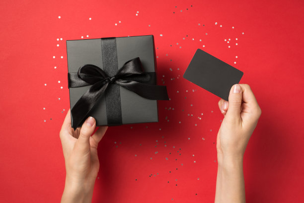 第一人称俯视图照片，女性手拿黑色塑料卡和黑色礼盒与黑色蝴蝶结和亮片孤立的红色背景空白图片下载