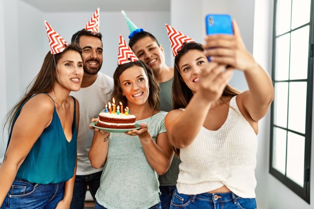 年轻的西班牙妇女微笑快乐，拿着生日蛋糕。一群朋友站在家里的智能手机旁自拍。图片下载