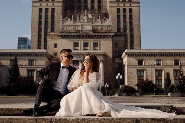 一对相爱的年轻夫妇的照片，身着婚纱的新娘和新郎坐在台阶上，背景是城市风景，戴着太阳镜，阳光明媚，幸福在一起。图片下载