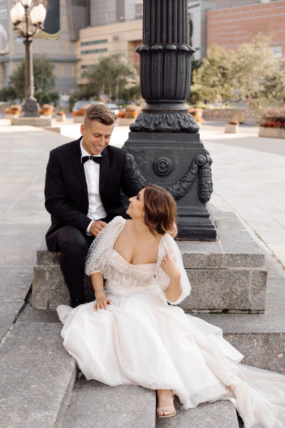 一对相爱的年轻情侣站在灯柱前，身着婚纱和旧城背景坐在楼梯上，共同祝新婚快乐。完整的照片图片下载