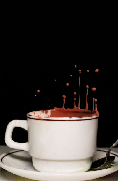 热巧克力从带有金属茶匙的白色杯碟中喷射出来，溅起的小滴在黑色背景下漂浮在半空中，聚焦前景，复制空间图片下载