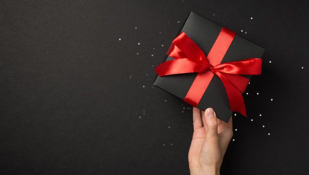 第一人称俯视照片的女性手拿黑色礼盒生动的红色蝴蝶结在闪亮的亮片与空白的孤立的黑色背景图片下载