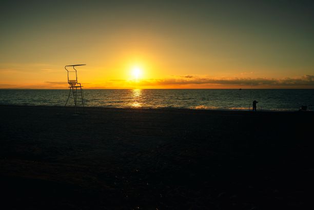 海滩上的日出。一个渔夫带着他的钓竿，而海滩守望者的椅子却空着。图片下载