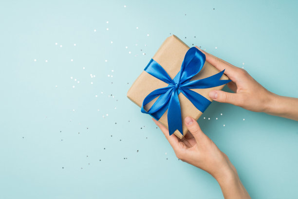 第一人称俯视图的照片，妇女的手赠送的手工纸礼品盒与蓝色缎带蝴蝶结在闪闪发光的亮片在孤立的淡蓝色背景与空白图片下载
