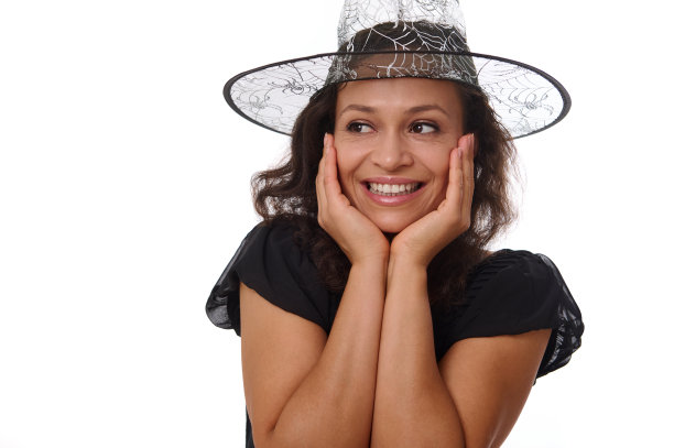 近肖像的一个漂亮的女人在女巫万圣节的帽子，手在脸上和可爱的微笑与美丽的牙齿微笑神秘地看着一边在白色背景的副本空间。图片下载