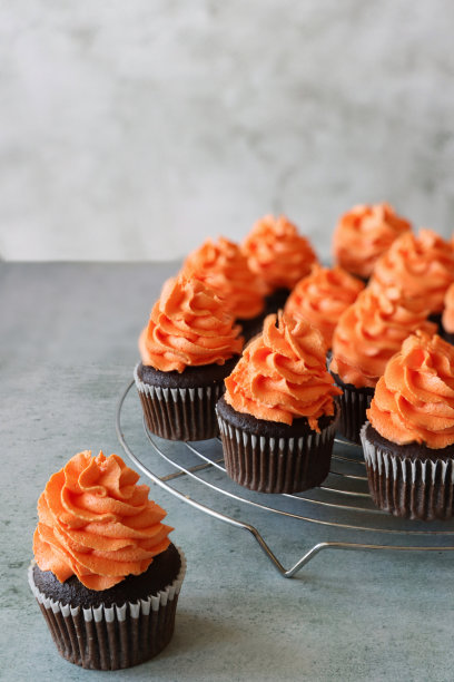 图片:一堆自制的万圣节巧克力纸杯蛋糕，纸盒放在金属冷却架上，顶部有橙色黄油糖霜，大理石效果的背景，聚焦前景图片下载
