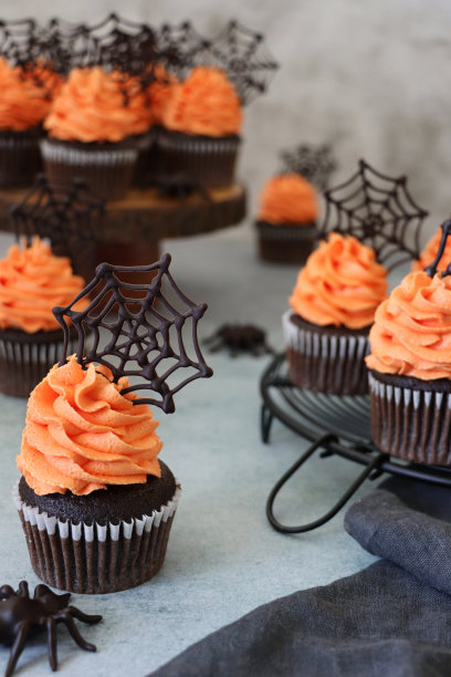 图片是批量自制的万圣节巧克力纸杯蛋糕，纸杯蛋糕盒放在木制蛋糕架和金属冷却架上，橙色黄油糖霜管道螺旋顶部有巧克力蜘蛛网，灰色棉布，塑料蜘蛛，大理石效果的背景图片下载