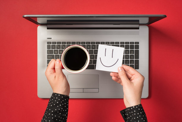 第一个人俯视图的照片，手握不干胶笔记纸画笑脸和一杯咖啡在开放的灰色笔记本电脑孤立的红色背景图片下载