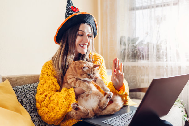 万圣节封锁。戴着女巫帽的女人拿着猫在笔记本电脑上视频聊天。假期在冠状病毒图片下载