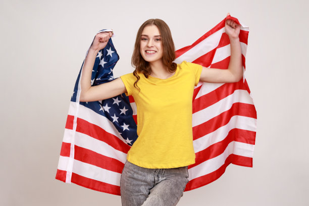 年轻的微笑女子，长相可爱，卷发，穿着休闲风格的黄色t恤，手举美国国旗，微笑着看着镜头。图片下载