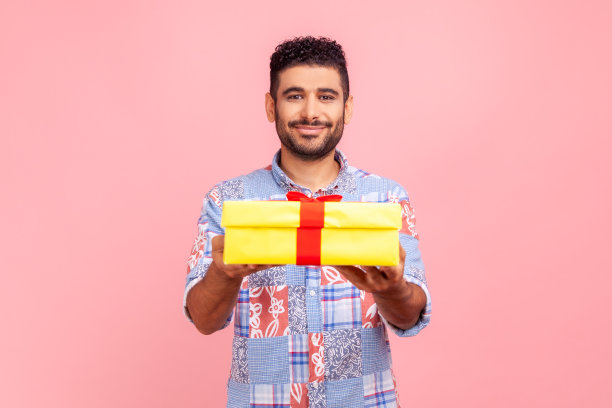 送给你！身穿蓝色休闲衬衫的快乐年轻男子将礼品盒送给镜头，脸上带着兴奋的微笑，在节日里问候和分享礼物图片下载