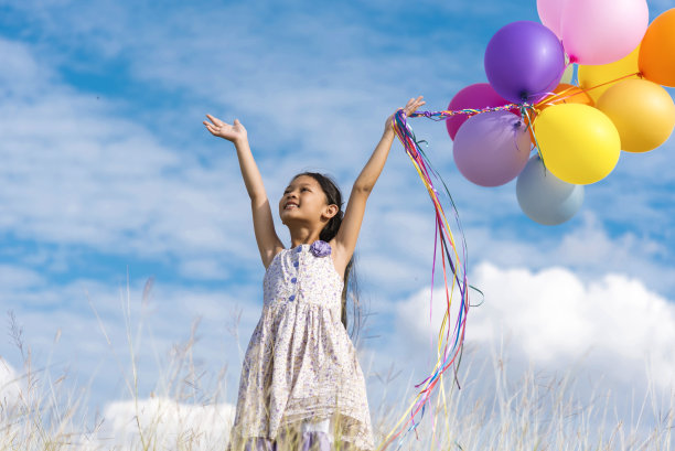 欢快可爱的女孩举着气球在绿色的草地、白云、蓝天上快乐地奔跑。手握着充满活力的气球在生日派对上玩耍快乐的时光，夏天在户外的阳光下图片下载
