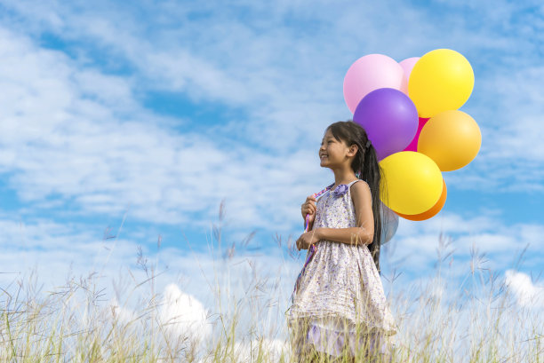 欢快可爱的女孩举着气球在绿色的草地、白云、蓝天上快乐地奔跑。手握着充满活力的气球在生日派对上玩耍快乐的时光，夏天在户外的阳光下图片下载