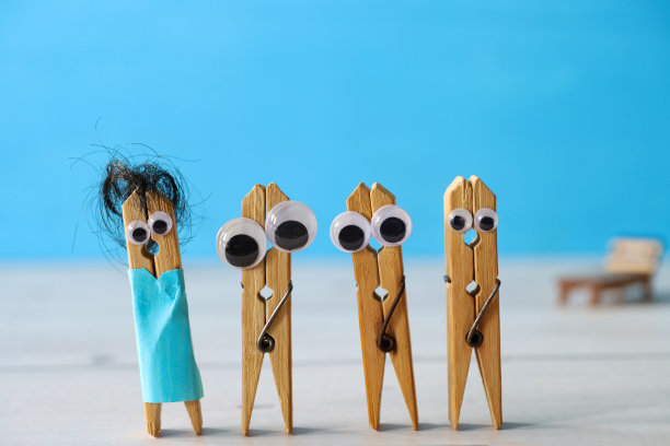 沙滩场景图片，由四个木制的衣服挂钩与大眼睛，一个挂钩与黑色的头发和纸板游泳衣，木制日光躺椅背后，蓝色背景，聚焦前景，幽默的概念图片下载