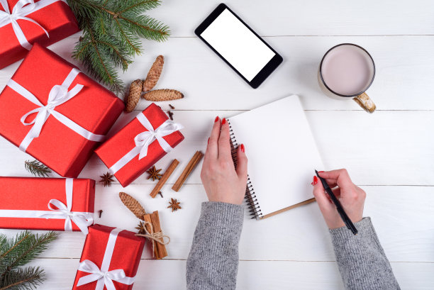 女人在笔记本上写下新年的目标(要做的事情)，桌子上有圣诞装饰品，红色的礼品盒，一杯可可或巧克力和有空白屏幕的手机，复印空间。俯视图图片下载