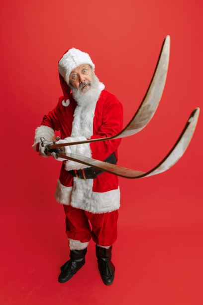 老人全身肖像穿着圣诞老人服装，手持滑雪板孤立在红色背景。冬季假期图片下载