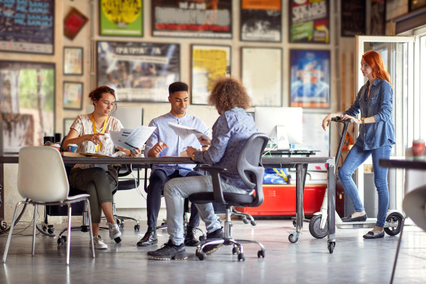 多民族的同事们一起工作，面带微笑，坐在办公桌前，他们的同事站在旁边的电动滑板车上图片下载