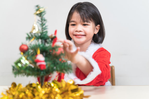 一个可爱的亚洲小女孩在圣诞夜穿着红色的圣诞老人服装装饰圣诞树。泰国圣诞假期概念库存照片图片下载