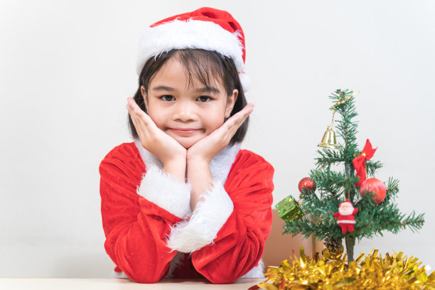 一个可爱的亚洲小女孩在圣诞夜穿着红色的圣诞老人服装装饰圣诞树。泰国圣诞假期概念库存照片图片下载