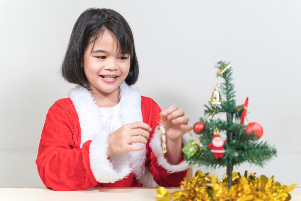 一个可爱的亚洲小女孩在圣诞夜穿着红色的圣诞老人服装装饰圣诞树。泰国圣诞假期概念库存照片图片下载