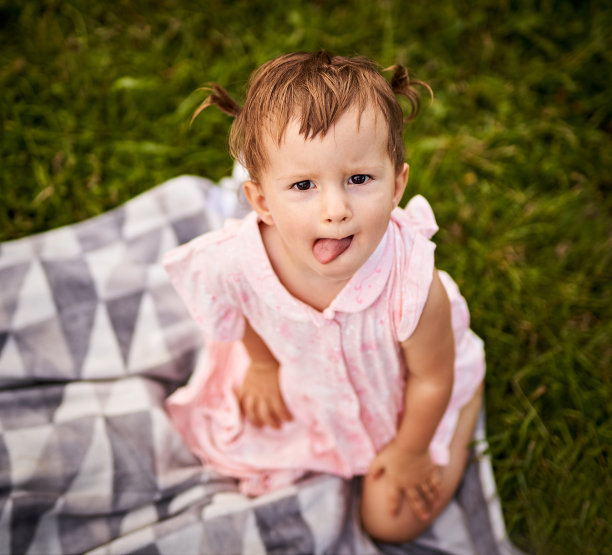 一个蹒跚学步的小女孩坐在野餐毯上，伸出舌头看着摄像机-小女孩享受生活和乐趣-家庭和童年户外概念在公园的俯视图图片下载