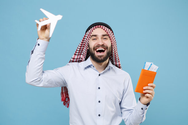 笑着的年轻阿拉伯穆斯林男子，留着胡须，穿着凯菲耶·卡iya戒指igal agal便服，孤立在淡蓝色的背景下。人们的宗教生活方式观念。持有护照、机票、飞机。图片下载