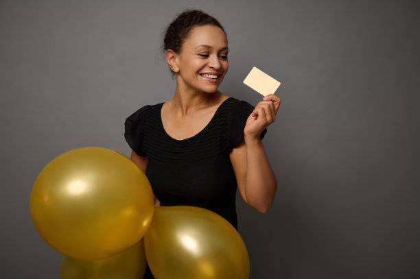 美丽的混血女子身穿黑色服装，在灰色的墙壁背景下微笑着，手里拿着气球和金色的空白信用卡。黑色星期五的概念与拷贝广告空间图片下载