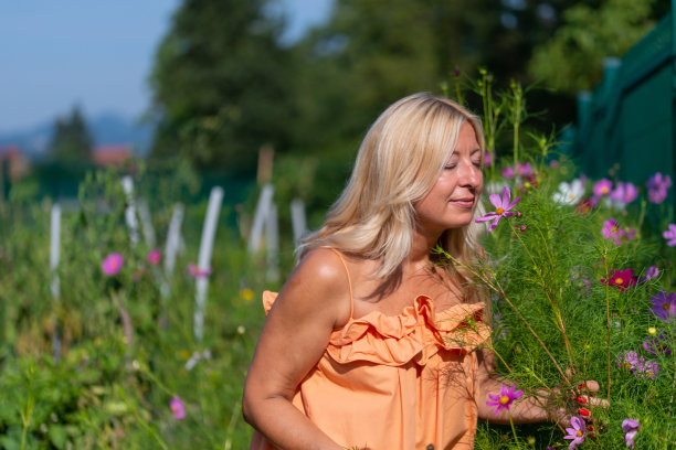 一个身穿橙色连衣裙的金发美女在菜园里收集、欣赏并闻着药草和盛开的花朵。图片下载