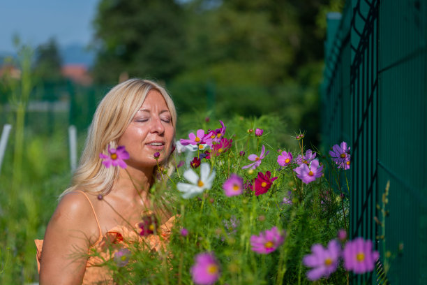 一个身穿橙色连衣裙的金发美女在菜园里收集、欣赏并闻着药草和盛开的花朵。图片下载