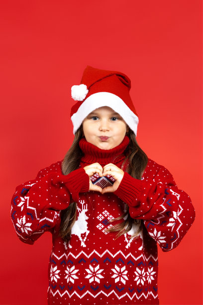淘气、顽皮的女孩的肖像，在红色针织圣诞毛衣与驯鹿和红色帽子显示心形标志的手指，孤立在红色背景图片下载