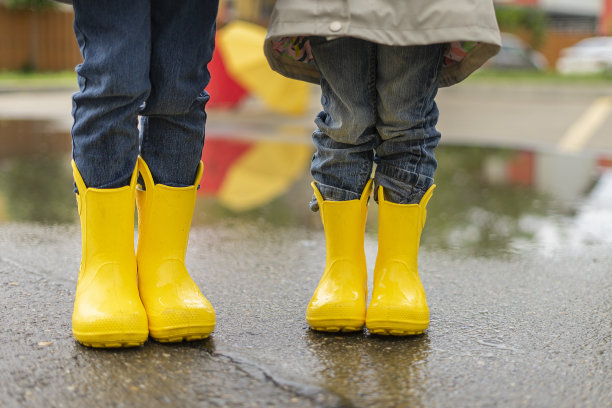 黄色的儿童胶靴在街上，旁边是雨伞作为背景。孩子跳进水坑里。雨后两个孩子的腿站在一起图片下载