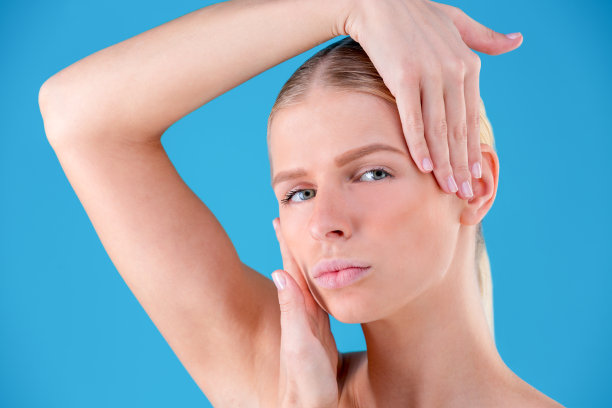 接近的肖像华丽的美丽和好看的年轻女人与无瑕的皮肤。她用手捂住孤立在蓝色背景上的脸颊图片下载