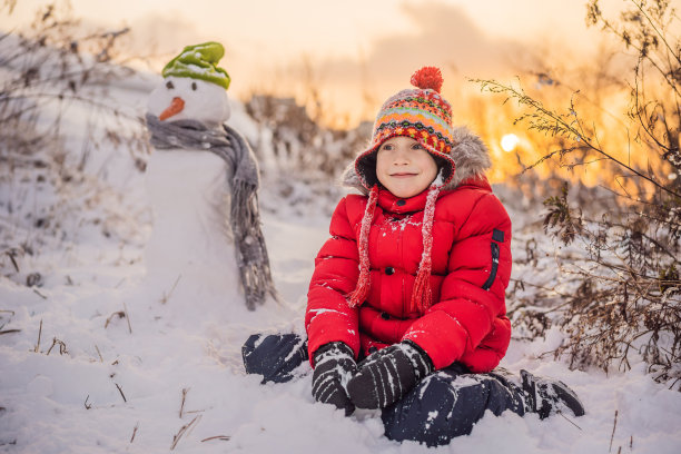 可爱的男孩穿着红色的冬衣堆雪人。冬季户外趣味概念图片下载
