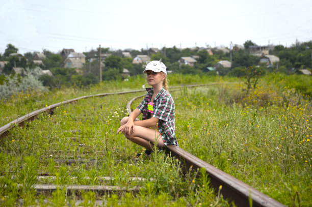 一个穿着格子衬衫、戴着帽子的女孩坐在一条废弃铁路的铁轨上图片下载