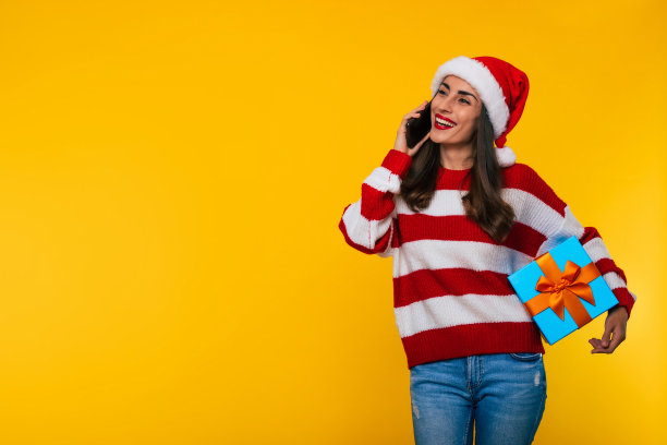 近距离照片，美丽兴奋微笑的女人与人交谈的圣诞节礼物盒和智能手机在黄色背景图片下载