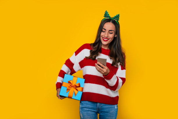 黄色背景下，带着圣诞礼盒和智能手机的美丽微笑女人正在使用设备的特写照片图片下载