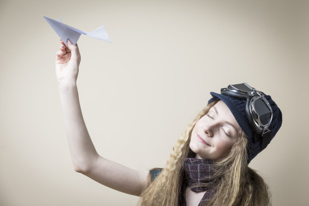 白种人金发少女在飞行眼镜和温暖的帽子与折纸飞机，而坐在旅行手提箱在米色背景。水平图像组成图片下载
