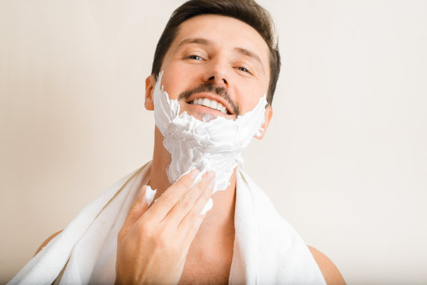 特写照片中英俊的正面男子肩膀上的毛巾涂抹保湿剃须泡沫，微笑的男子看着相机，做他的日常剃须，皮肤护理，男性美容图片下载