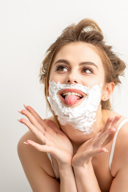 一幅美丽的年轻白人女子的肖像面带微笑，脸上抹着剃须泡沫，在白色背景下伸出舌头。图片下载
