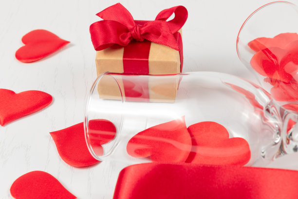 两个香槟杯，一个装着礼物的盒子，一条红色的丝带和散落在白色木桌上的一颗心图片下载