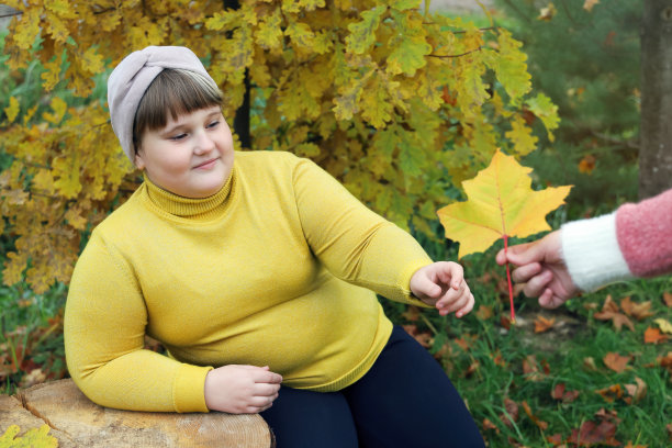 肥胖微笑的女孩坐在公园里的秋树旁边，从另一棵黄色的枫叶图片下载