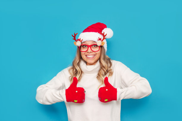 一个年轻的女人穿着白色温暖的毛衣，戴着红色的圣诞老人帽，用她的手做着大拇指朝上的手势。圣诞节和新年的概念图片下载