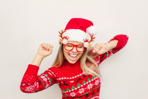 一位身穿马鹿毛衣、头戴圣诞老人帽子的年轻女子高兴地举起双手。圣诞节和新年的概念图片下载