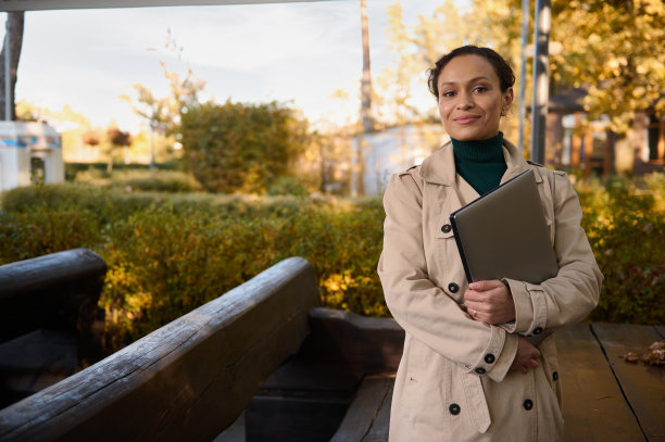 自信的肖像迷人的非洲商业女性在休闲米色风衣和绿色套头衫看着相机，交叉双臂拿着笔记本电脑在秋天的自然背景图片下载
