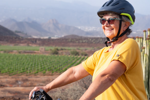 老年妇女喜欢骑自行车户外远足和健康的生活方式。戴着运动头盔的老年妇女在葡萄园和山区的乡村微笑图片下载