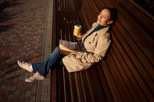 公园里的一张木凳上，一位美丽宁静的年轻女子背靠在椅背上，拿着一个一次性纸杯，里面装着热饮和一本书，远离了城市的喧嚣，享受着阳光明媚的一天图片下载