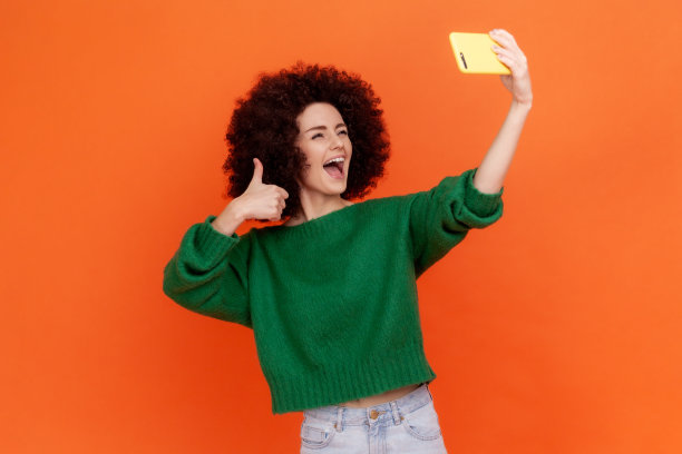 一名留着非洲式发型、身穿绿色休闲风格毛衣的兴奋快乐的女子在粉丝中直播，并对着镜头竖起大拇指。图片下载