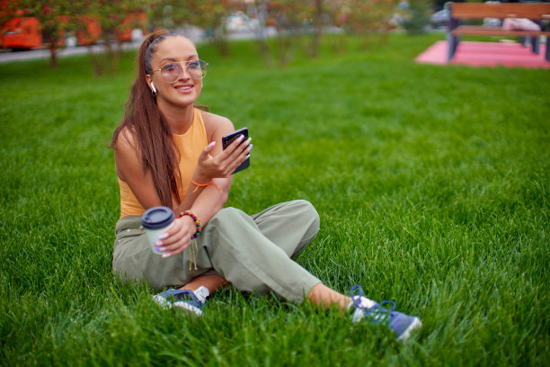 快乐的年轻女孩戴着耳机，用手机应用听音乐或播客，在户外草坪上放松图片下载