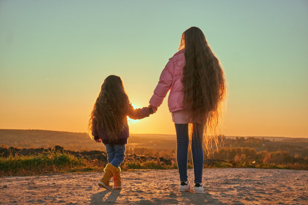 长发飘逸的两姐妹手牵着手站在那里，向着地平线。看日落。秋天的天气是一条土路。夕阳的光线穿透松散的头发图片下载