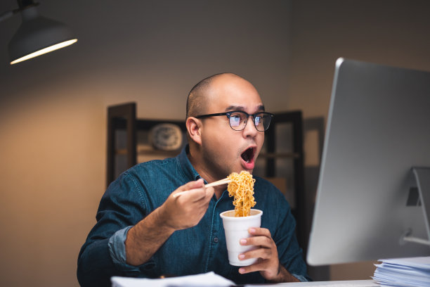 年轻的亚洲自由职业者工作到深夜。晚上他很饿，吃着热腾腾的方便面，在黑暗的办公室里用电脑工作。图片下载